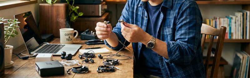 自宅デスクで、数種類のイヤホンを並べ、それぞれの形やマイクの位置を興味深そうに観察している男性